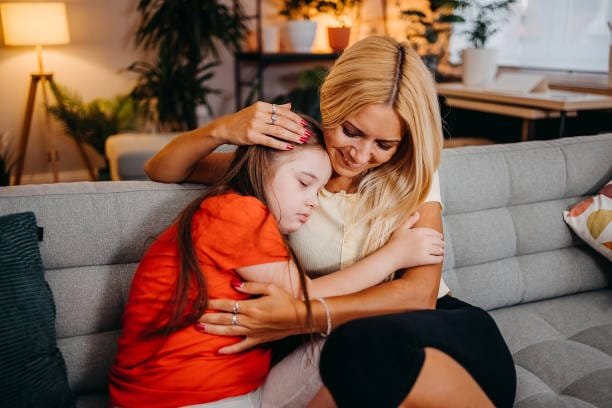 Young mother embracing her disabled child while they sitting on sofa in the room